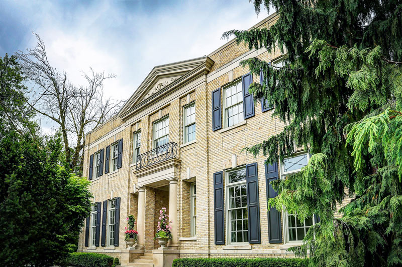 A stately yellow-brick luxury home with navy shutters surrounded by mature trees